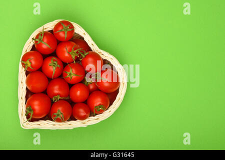 Tomates cerises fraîches mûr rouge en forme de coeur dans un petit panier en osier vert sur fond de papier Banque D'Images