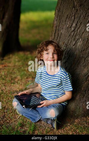 Le garçon de 8-9 ans est assis appuyé contre un arbre et détient le comprimé dans la main.avec une blonde Laddie cheveux bouclés ressemble à un appareil photo Banque D'Images