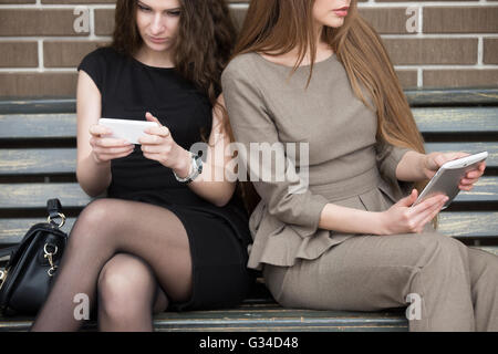 Deux belles jeunes femmes assises côte à côte sur le banc sans parler et de leurs propres appareils. Accro du téléphone Banque D'Images