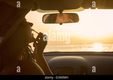 Woman applying lipstick et rétroviseur à l'aide de voiture au coucher du soleil Banque D'Images