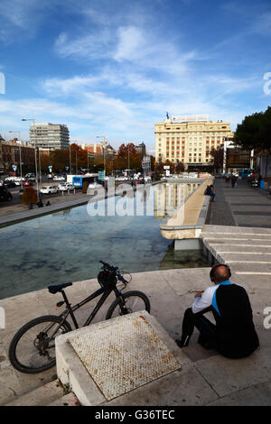 Homme portant des écouteurs assis à côté de la bicyclette à Jardines del Descubrimiento, Plaza de Colon, Madrid, Espagne. Hôtel Fénix Gran Meliá en arrière-plan. Banque D'Images