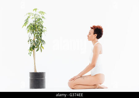 Calme african american woman with eyes closed assis près de petit arbre en pot sur fond blanc Banque D'Images