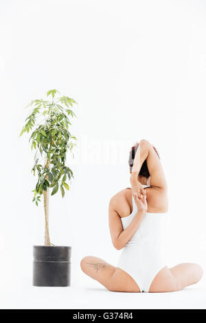 Vue arrière de pretty african american woman stretching arms et pratiquant le yoga près de petit arbre en pot sur fond blanc backgroun Banque D'Images