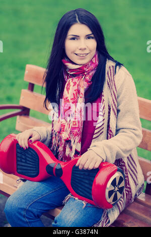 Jeune fille brune holding electric moderne rouge mini hover board scooter dans les mains tout en restant assis sur le banc de parc. Nouvelles populaires electric city transport. Fille porte des vêtements de style Boho de tendances. Banque D'Images