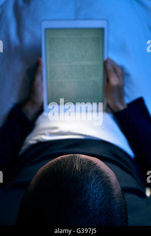 Un jeune homme de race blanche au lit lit un e-book dans son ordinateur, tablette de nuit Banque D'Images
