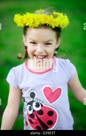 La petite fille dans une couronne de pissenlits jaunes. Le cheerful girl sur un fond vert. Les cheveux sont tressés en petites nattes. O Banque D'Images