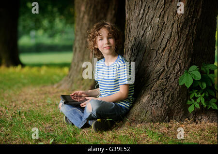 Le garçon de 8-9 ans est assis appuyé contre un arbre et détient le comprimé dans la main. Le petit gars avec une blonde cheveux bouclés ressemble je Banque D'Images