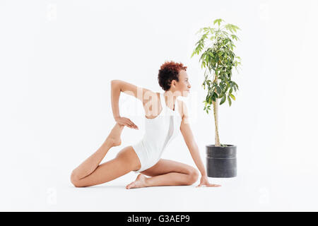 Belle jeune femme afro-américaine de l'exercice et la pratique du yoga près de petit arbre en pot sur fond blanc Banque D'Images