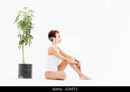 African American pacifique jeune femme assise près de petit arbre en pot sur fond blanc Banque D'Images