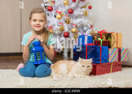 Fille de sept ans est assis avec un chat sous l'arbre de Noël avec des cadeaux et souriant joyeusement Banque D'Images