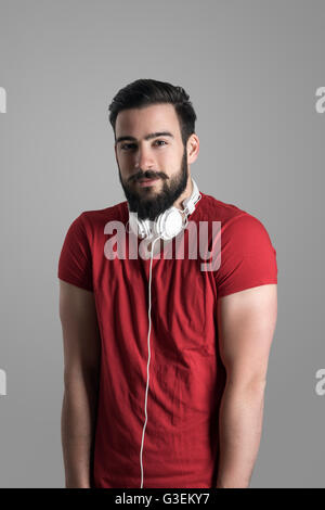 Jeune homme barbu avec un casque sur le cou qui posent avec les mains dans les poches et les épaules relevées jusqu Banque D'Images