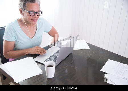 Senior woman sitting at table, en utilisant un ordinateur portable, sur la table de la paperasserie Banque D'Images