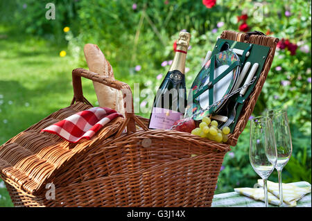 Bouteille de vin rosé mousseux anglais et panier pique-nique en osier avec baguette et raisins dans un jardin anglais fleuri ensoleillé Banque D'Images