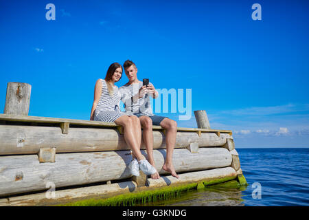 Couple sitting on wooden pier, taking self portrait using smartphone Banque D'Images