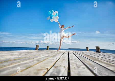 Jeune femme dansant sur jetée en bois, holding bunch of balloons Banque D'Images