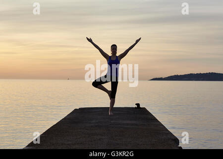 Jeune femme debout sur la jetée, en position de yoga, au coucher du soleil Banque D'Images