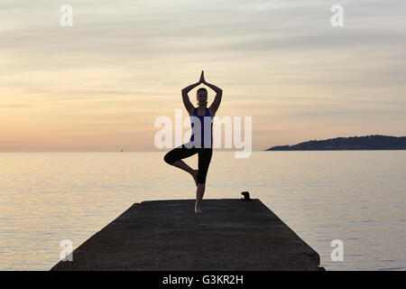 Jeune femme debout sur la jetée, en position de yoga, au coucher du soleil Banque D'Images