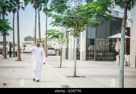 Homme portant des vêtements traditionnels à marcher le long street talking on smartphone, Dubai, Émirats Arabes Unis Banque D'Images