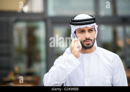 Homme portant dishdasha marcher le long street talking on smartphone, Dubai, Émirats Arabes Unis Banque D'Images