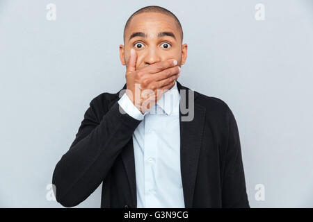 Beau jeune homme africain couvrant la bouche à part isolé sur fond gris Banque D'Images