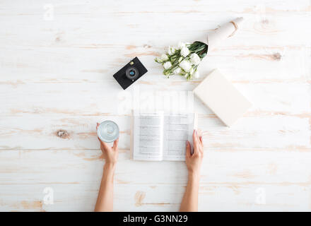 Les mains des femmes plus de tourner les pages d'un livre avec un verre d'eau, des fleurs et de la caméra sur le bureau en bois Banque D'Images