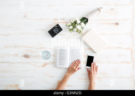 Les mains des femmes plus de tourner des pages de livre et holding smartphone sur le bureau en bois Banque D'Images