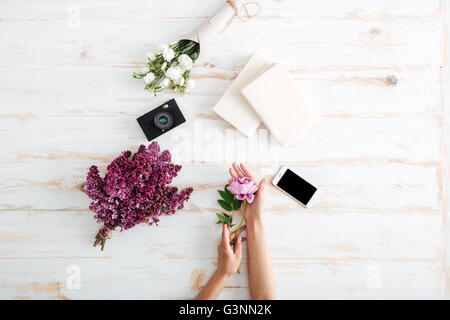 Les mains des femmes avec des fleurs de pivoine, livres, photo, verre d'eau et lilas bouquet sur le bureau en bois Banque D'Images