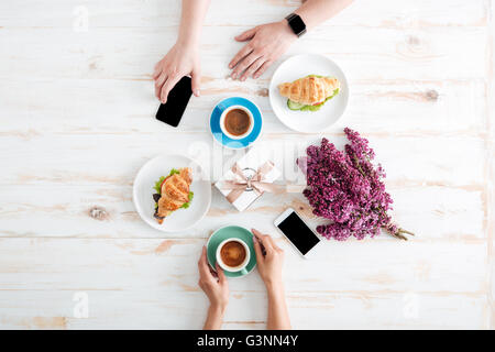 Mains de jeune couple de boire du café avec des croissants et à l'aide d'écran vide smartphones sur table en bois Banque D'Images