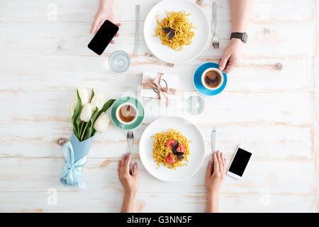 Mains de jeune couple eating pasta et à l'aide d'écran vide smartphones sur table en bois avec tasses de café, boîte-cadeau et verres Banque D'Images