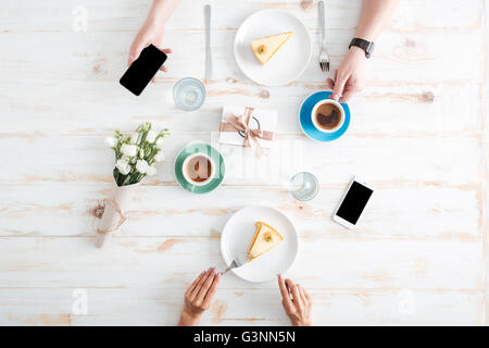 Mains de jeune couple de manger des gâteaux et à l'aide d'écran vide smartphones sur table en bois avec tasses de café fort, présent et rose ... Banque D'Images