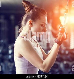 Jeune femme slim en pile faire sur le câble de la machine en salle de sport dans le coucher du soleil de poutres apparentes. Athletic Girl triceps de formation dans un centre de remise en forme Banque D'Images