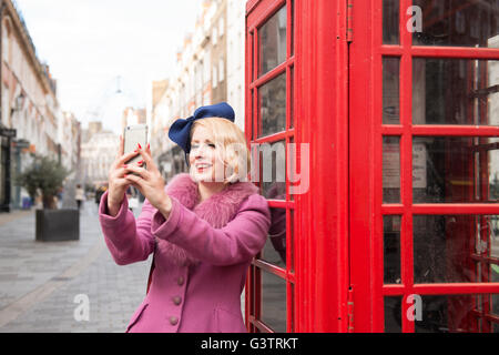 Une élégante jeune femme vêtue de vêtements de style années 30 en tenant un téléphone traditionnel selfies en dehors d'une buvette sur une rue de Londres. Banque D'Images