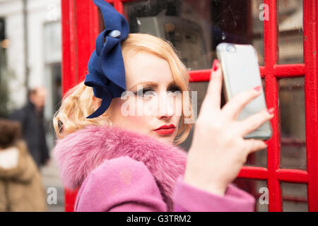 Une élégante jeune femme vêtue de vêtements de style années 30 en tenant un téléphone traditionnel selfies en dehors d'une buvette sur une rue de Londres. Banque D'Images