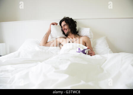 Un jeune homme aux cheveux longs couché dans un lit avec une tasse de thé à la recherche heureux. Banque D'Images