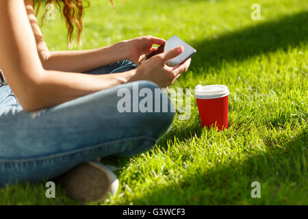 Jeune femme avec du papier jetable rouge tasse de café à l'aide de tablet pc sur une pelouse verte dans un parc Banque D'Images