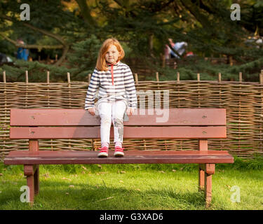 Assis sur un banc. Une jeune fille aux cheveux rouges se trouve sur l'arrière d'un banc de parc face à l'appareil photo. Banque D'Images