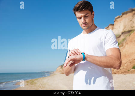 Beau jeune sportsman et permanent à l'aide de smart watch sur la plage Banque D'Images