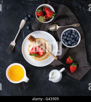 Des crêpes avec des fraises et des bleuets, des bols de miel et pot de lait sur serviette textile sur fond texturé noir. Bre Banque D'Images