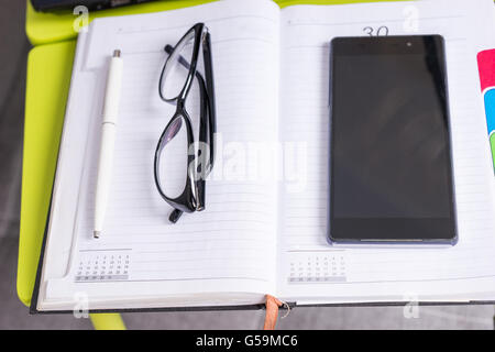 Se concentrer sur des verres élégants allongé sur la table ordinateur portable à proximité d'une page vierge d'un journal intime avec un stylo pour prendre des rendez-vous, l'organisation d'un calendrier et d'un téléphone mobile. Banque D'Images