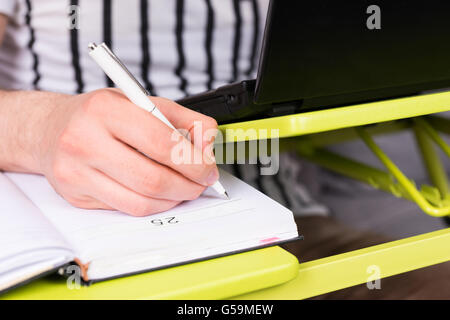 L'accent sur la main de l'homme écrit et de faire des nominations dans journal étendu sur la table ordinateur portable faire son travail d'accueil assis sur un canapé la tenue d'un stylo. Banque D'Images