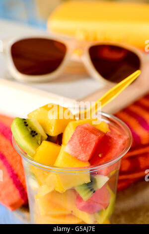 Libre d'un verre en plastique avec une salade de fruits avec une paire de lunettes de soleil, un livre et une serviette de plage colorée à l'arrière-plan Banque D'Images