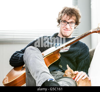 Smiling young man wearing glasses assis sur une chaise à la maison avec sa guitare et son chien de compagnie, un petit chihuahua reposant sa tête sur Banque D'Images