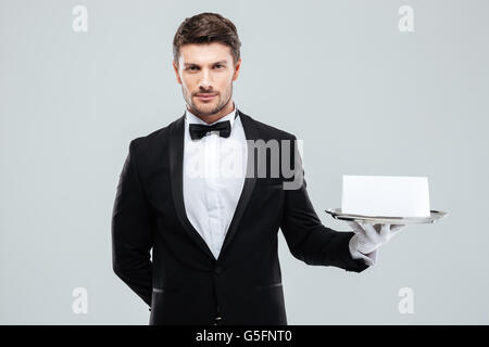 Young butler in tuxedo et gants holding tray with carte vierge Banque D'Images