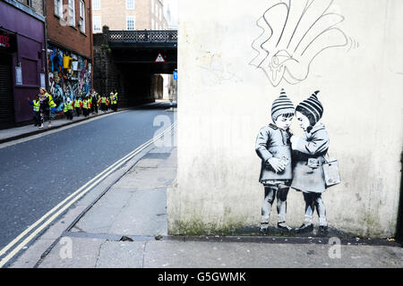 Un groupe de jeunes écoliers, portant des tabards réfléchissants, passent devant une fresque graffiti de style Banksy, de deux enfants chuchotant, sur un mur de bâtiment. Banque D'Images