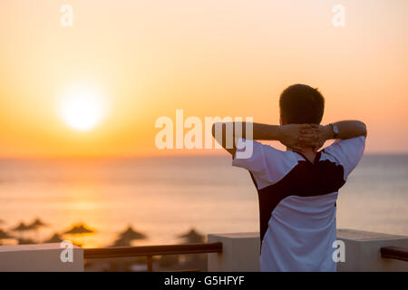 Jeune homme debout sur le balcon à carefree posent avec les mains derrière la tête, profitant de belles couleurs du lever ou du coucher du soleil sur la mer Banque D'Images