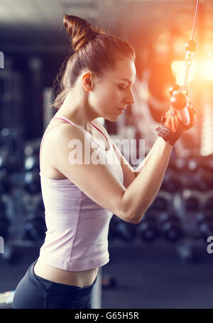 Jeune femme slim en pile faire sur le câble de la machine en salle de sport dans le coucher du soleil de poutres apparentes. Athletic Girl triceps de formation dans un centre de remise en forme Banque D'Images