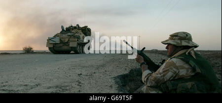 Un soldat britannique de l'Artillerie royale du 29 Commando Regiment est en service de garde, tandis qu'un char de Scimitar des gardes de dragons du Queens passe à côté, et qu'un feu de pétrole brûle à l'horizon dans le sud de l'Irak.Alors que le conflit irakien entre dans sa deuxième semaine. Banque D'Images