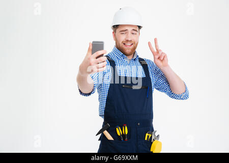 Smiling handsome young builder montrant signe de la paix et de faire avec un téléphone mobile selfies Banque D'Images