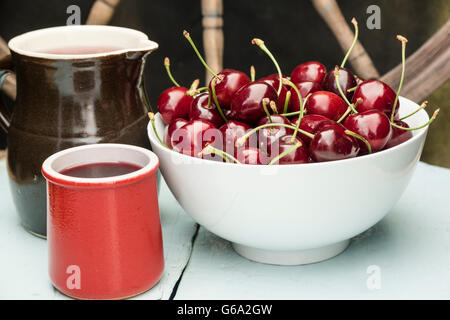 Les cerises dans un bol blanc, debout sur une table en bois bleu avec du jus dans une cruche d'argile et de la cuvette, avec roue en bois en arrière-plan Banque D'Images