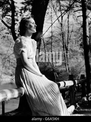 Années 1930 Années 1940 PORTRAIT THOUGHTFUL WOMAN SITTING ON FENCE SOUS LES ARBRES ombragés Banque D'Images